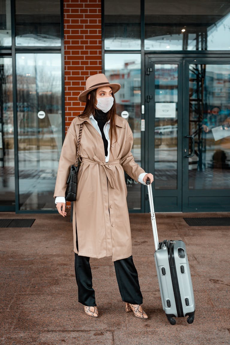 Woman standing outside with a suitcase, wearing a face mask and beige trench coat, ready for travel.
