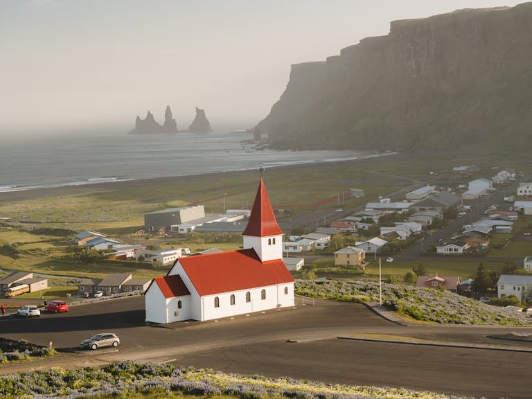 Aerial view of the iconic red-roofed Vik Church with Reynisdrangar sea stacks along the stunning Icelandic coastline.