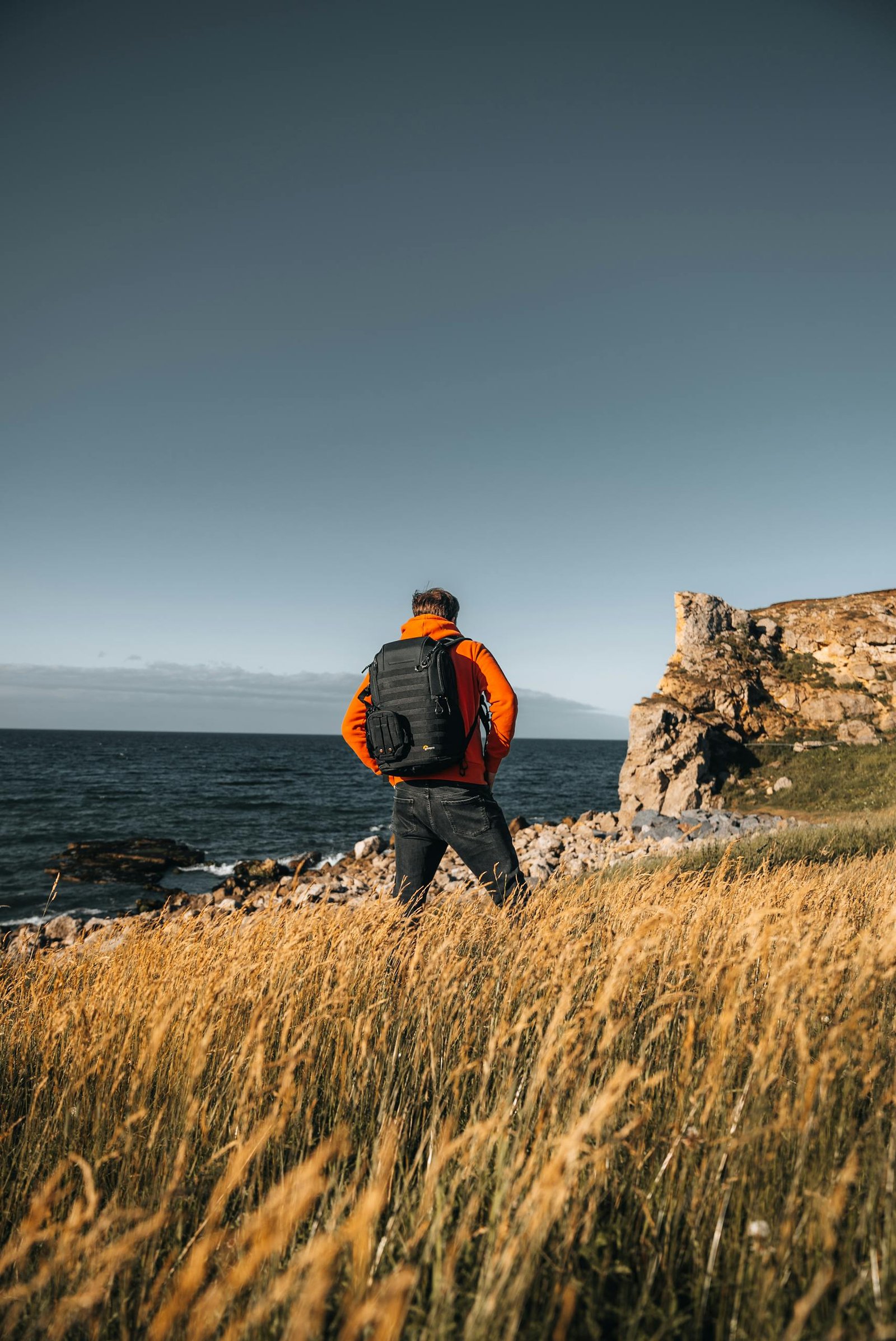A man in an orange jacket stands with a backpack, facing the ocean on a rocky coast under a clear sky.