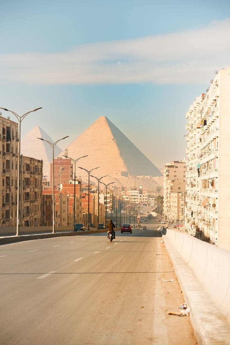 A street view in Cairo leading to the iconic Giza pyramids, with city buildings and traffic.