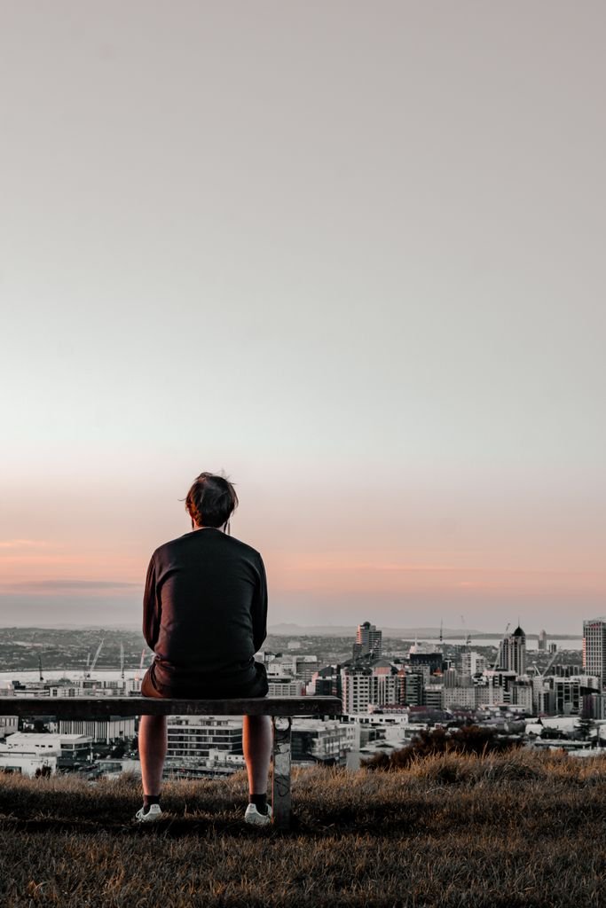 Man sits on a bench overlooking Auckland skyline at sunset, capturing solitude and reflection.