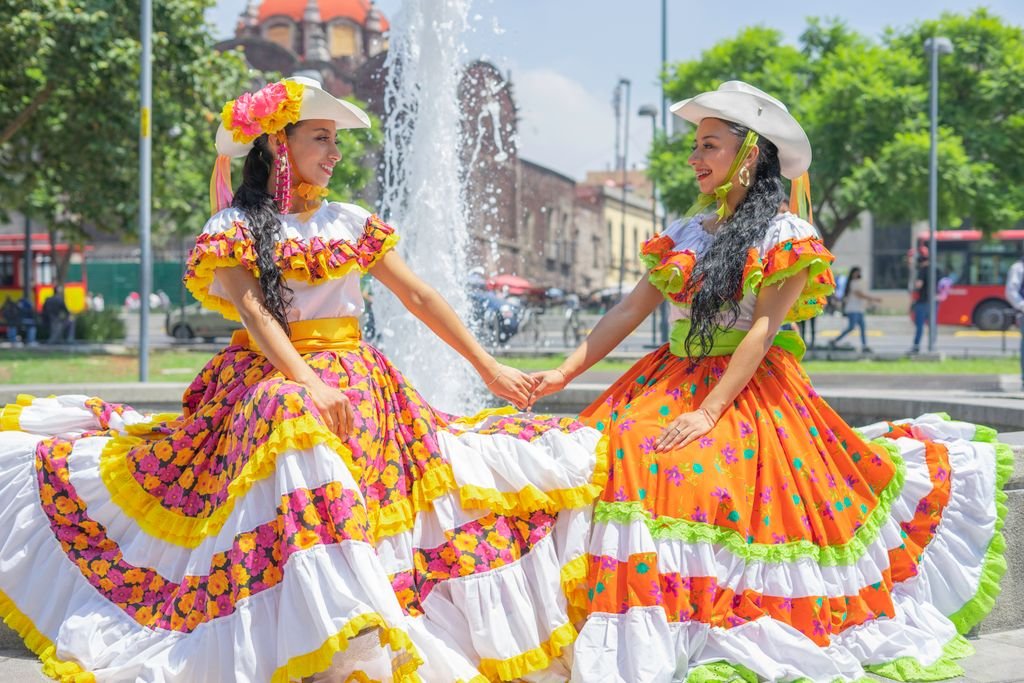 Two women in traditional Mexican dresses sit by a fountain, showcasing vibrant folkloric fashion.