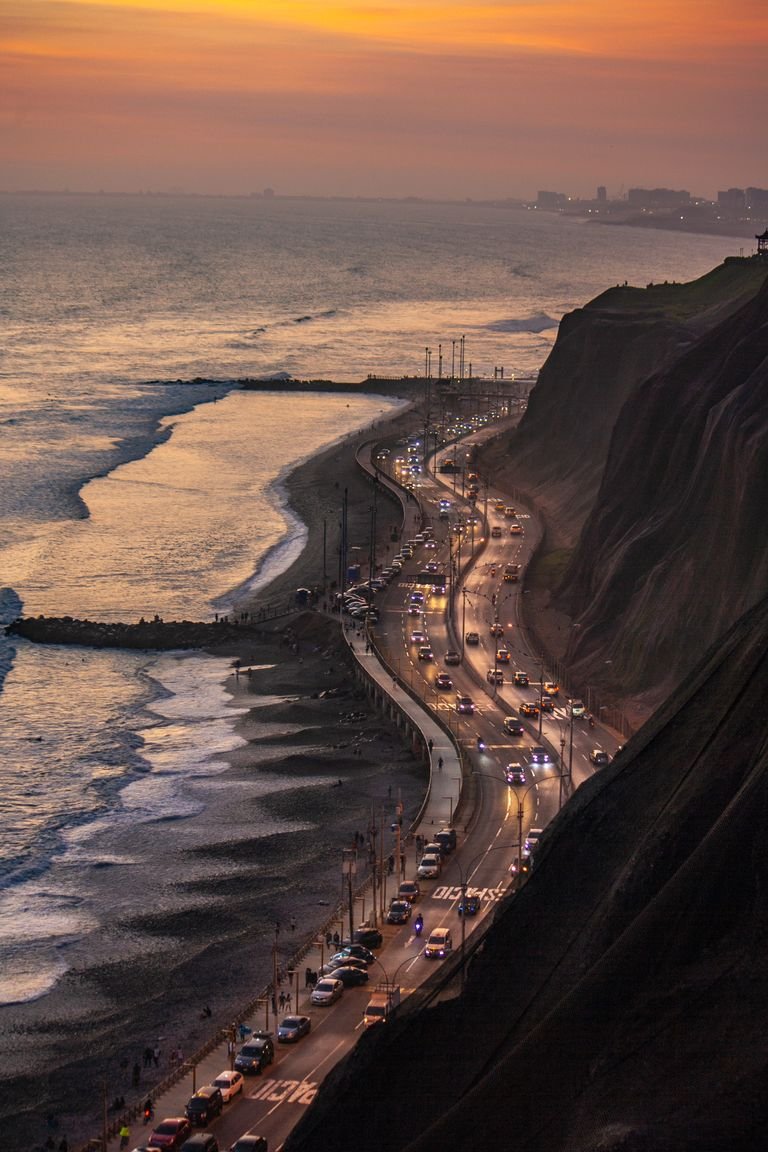A breathtaking view of a coastal road with traffic by a rugged shore at sunset, highlighting the ocean's beauty.
