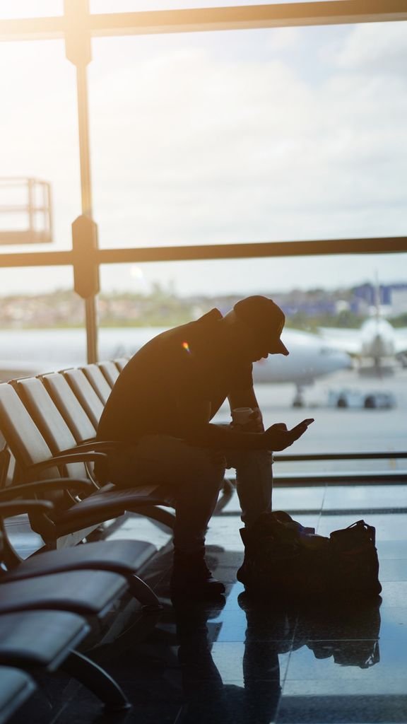 Silhouette of a man using a smartphone in an airport waiting area with airplanes visible outside.